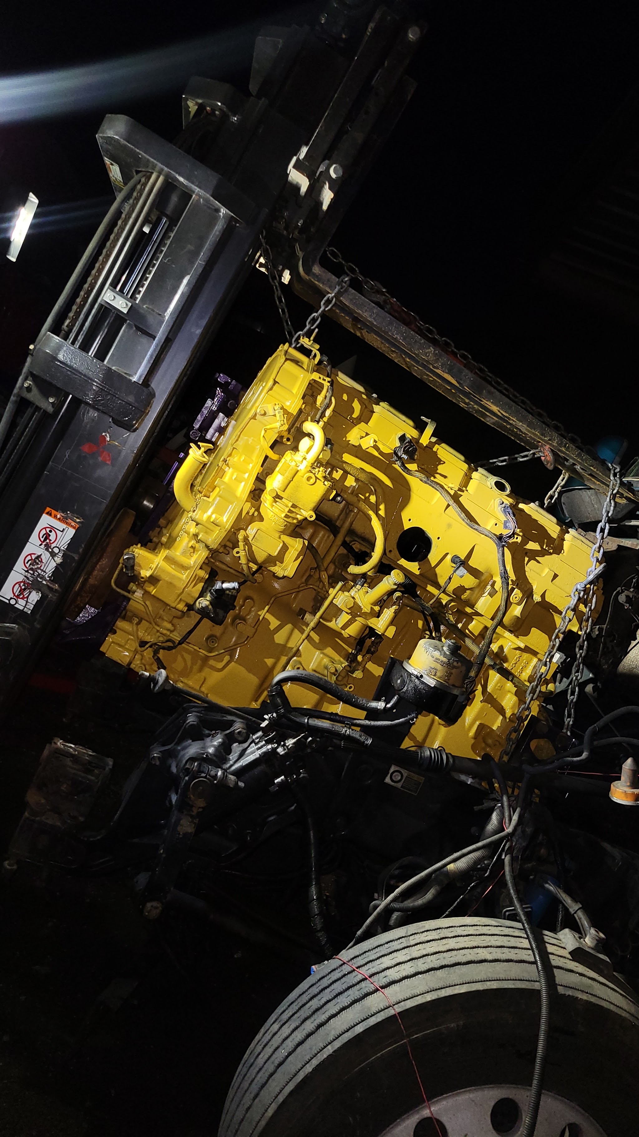 A freshly painted yellow truck engine hanging from a forklift chain at night
